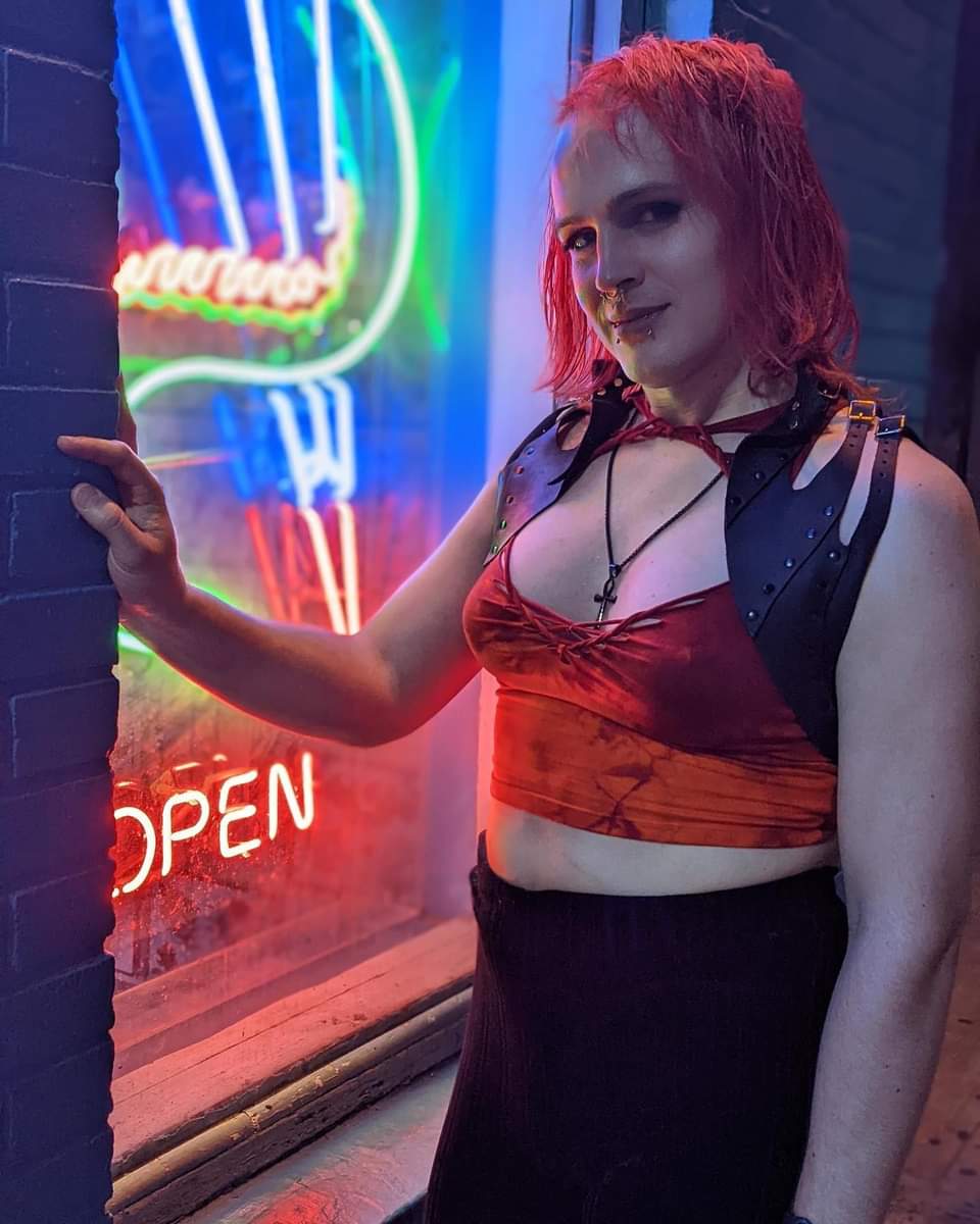 Futuristic headshot of Juno, who has bright red hair, a septum piercing, and symmetric vertical labret lip piercings. They are wearing a red and orange crop top, a leather shrug, an ankh necklace, and a black skirt. Juno is partially backlit by a neon display in a window which includes the word 'open.'